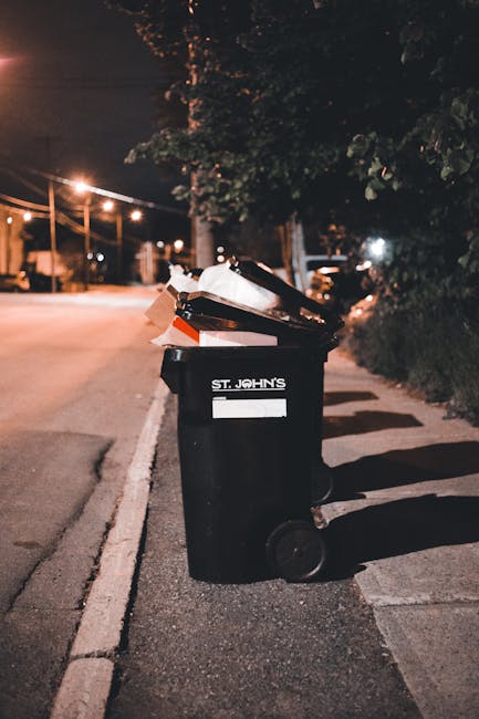 A pile of discarded rubbish on a gravel surface, including black plastic garbage bags, a broken beige car seat with fabric upholstery, a yellow plastic container, and various miscellaneous waste items