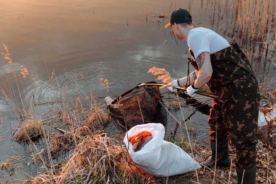 A person wearing a black cap, white t-shirt, camouflage overalls, and gloves stands at the edge of a wetland pond during daylight, using a net attached to a long handle to remove floating debris and a